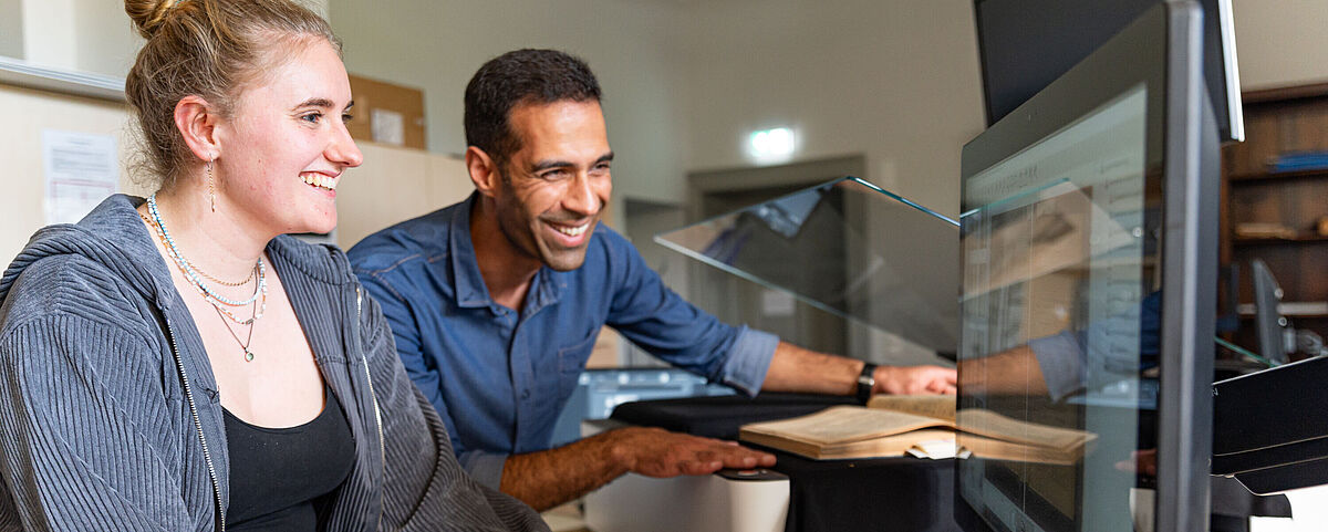 Arbeitgebermarketing Bibliotheken Eine Frau und ein Mann scannen gemeinsam etwas ein und schauen lächelnd auf einen Bildschirm.