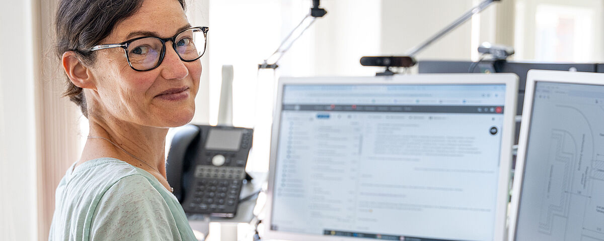 Arbeitgebermarketing Dezernat Planung und Technik Eine Frau mittleren Alters sitzt vor einem PC mit großem Monitor und lächelt freundlich in die Kamera.