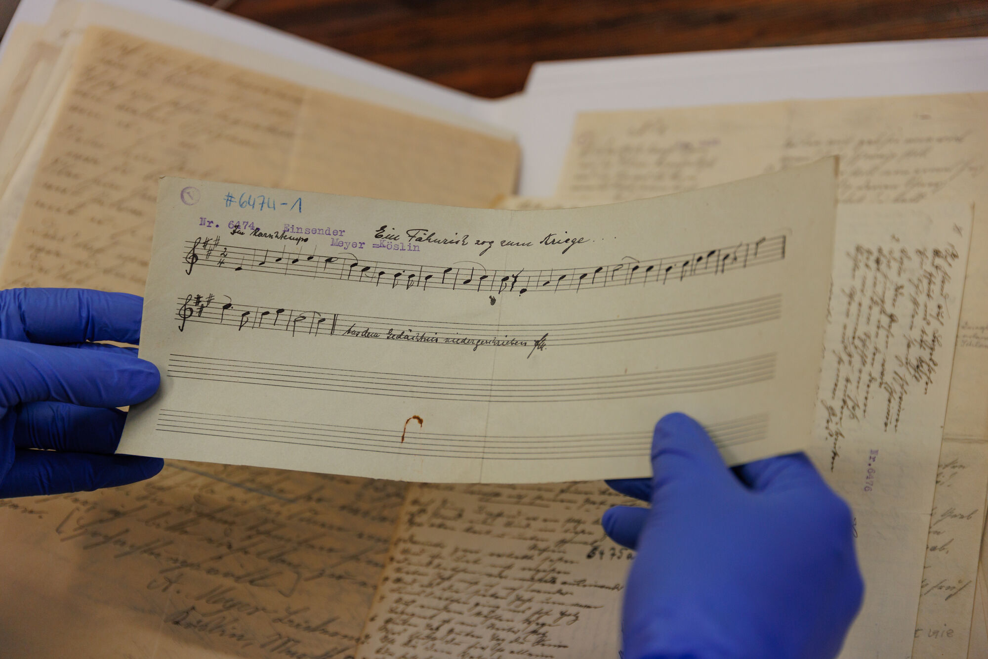 Symbolbild Pommersches Volksliedarchiv Ein Paar Hände in blauen Handschuhen hält ein handgeschriebenes Musikblatt mit Notenzeilen und einer Beschriftung, im Hintergrund liegen weitere handschriftliche Dokumente.