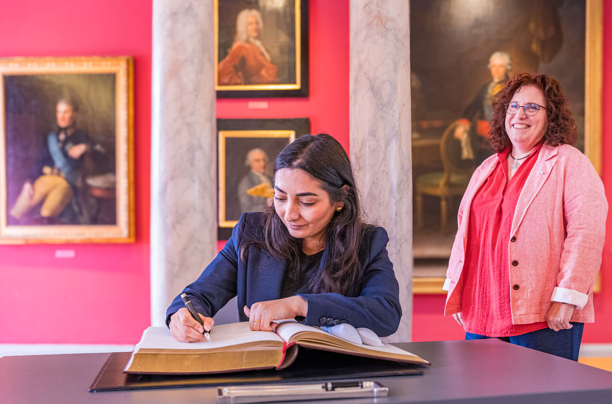 Die Bundesministerin für wirtschaftliche Zusammenarbeit und Entwicklung, Reem Alabali Radovan, trägt sich ins Ehrenbuch der Universität ein. Bundesministerin für wirtschaftliche Zusammenarbeit und Entwicklung, Reem Alabali Radovan, trägt sich ins Ehrenbuch der Universität ein. Sie sitz an einem Tisch in der Aula. Die Rektorin beobachtet sie dabei.