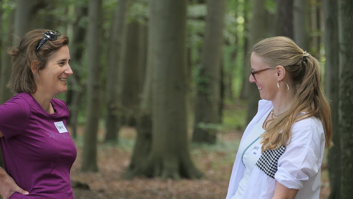 Zwei Frauen stehen sich gegenüber und unterhalten sich. Die Umgebung ist ein Wald.