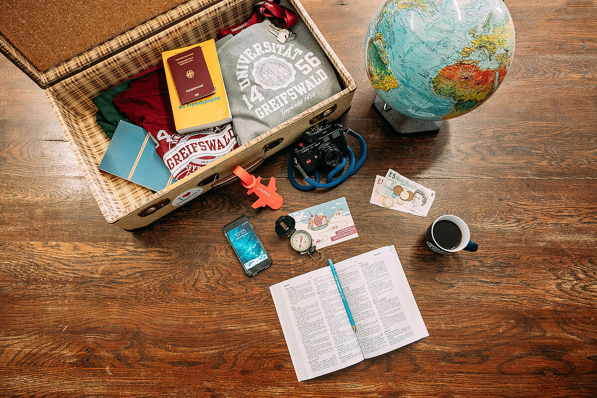 Koffer / Stillleben / International Office Auf einem Tisch liegt ein geöffneter Koffer, in ihm liegen ein grauer Hoodie, ein rotes T-Shirt, ein gelbes Wörterbuch, ein Schlüssel mit rotem Schlüsselband sowie ein Pass. Neben dem Koffer steht ein Globus. Vor dem Koffer liegt eine Kamera, ein Smartphone, ein Kompass sowie Geld und ein aufgeschlagenes Wörterbuch. Daneben steht eine Tasse mit Kaffee.