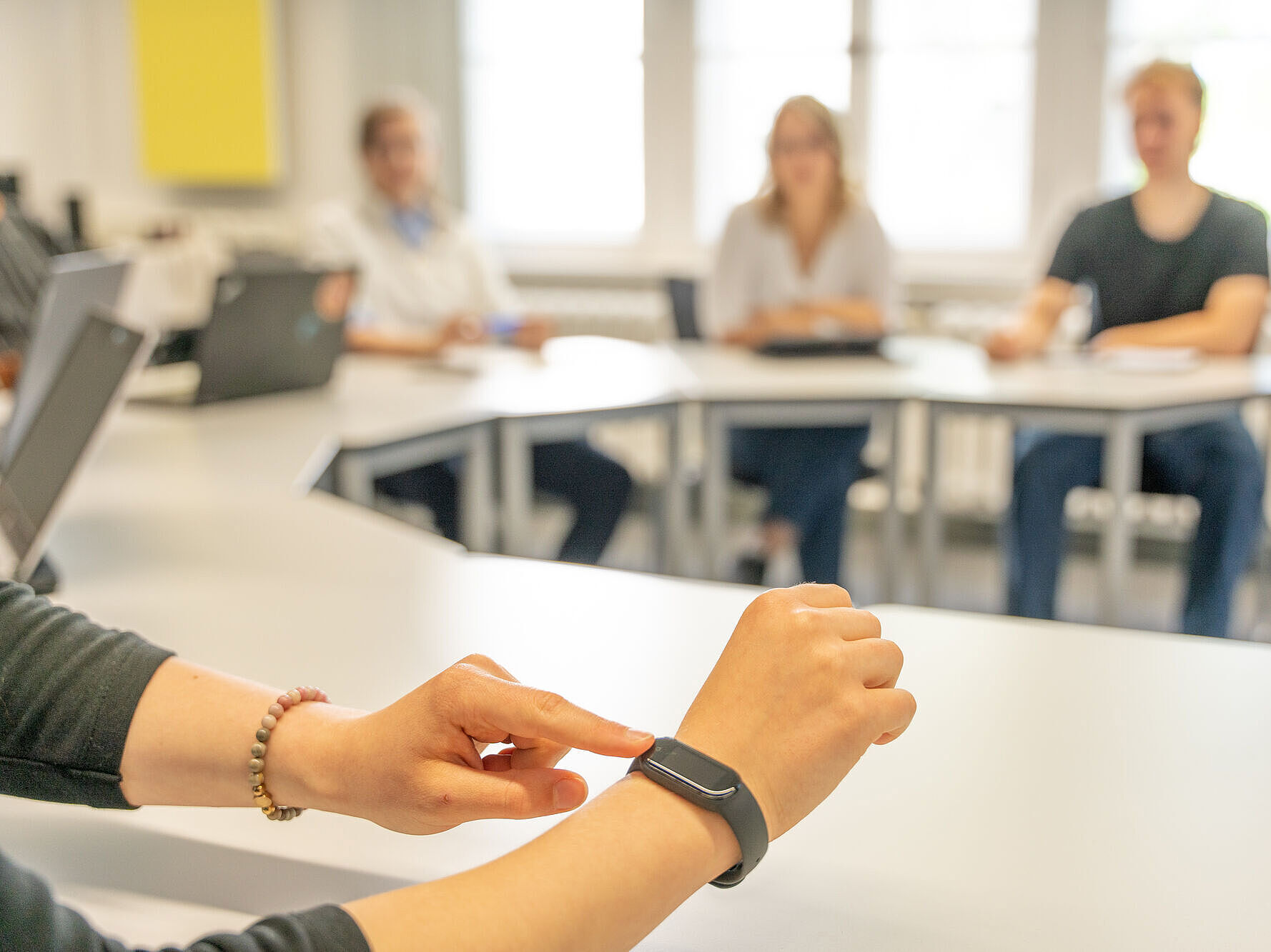 Im Vordergrund wird eine Smartwatch an einem Handgelenk bedient, während im Hintergrund unscharf vier Personen an Tischen in einem hellen Raum sitzen.