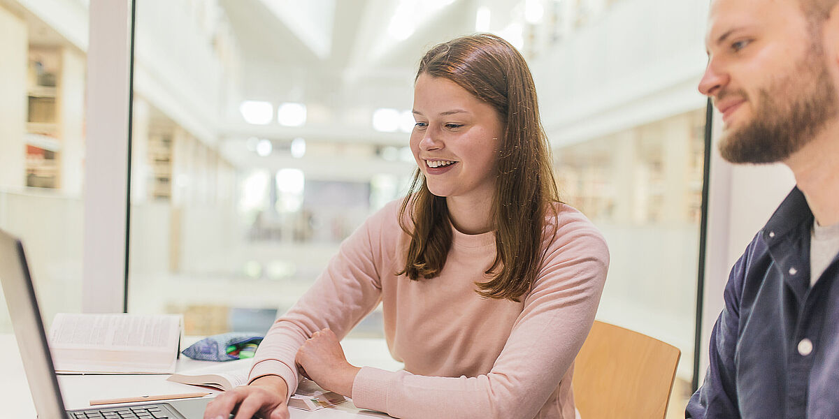 Mit Notebook in der Bibliothek Zwei Personen sitzen mit einem Notebook in der Bibliothek.