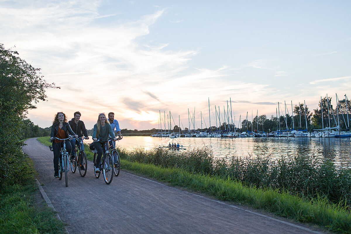 Heimathafen-Kampagne Eine Gruppe Radfahrende auf dem Treidelpfad am Ryck. Rechts im Bild sind Boote im Hafen sichtbar.