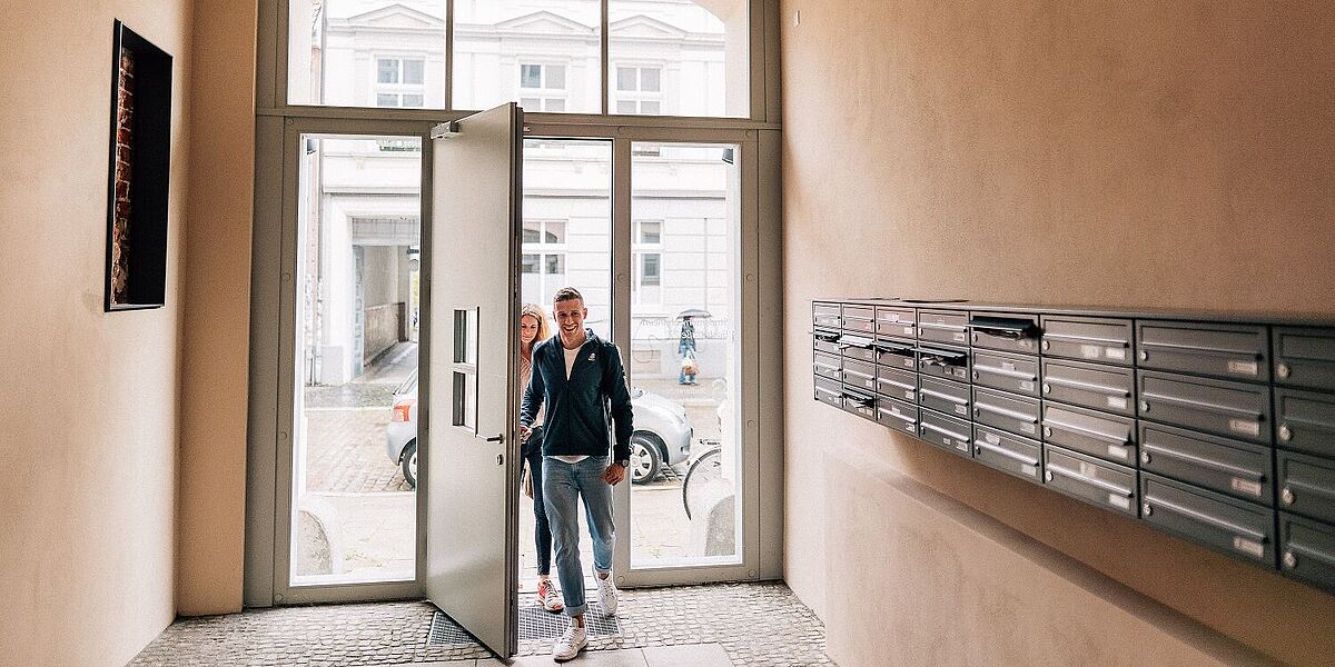 Wohnheim Studierendenwerk Zwei Studierende betreten das helle Treppenhaus von außen. Rechts sind Briefkästen an der Wand angebracht.