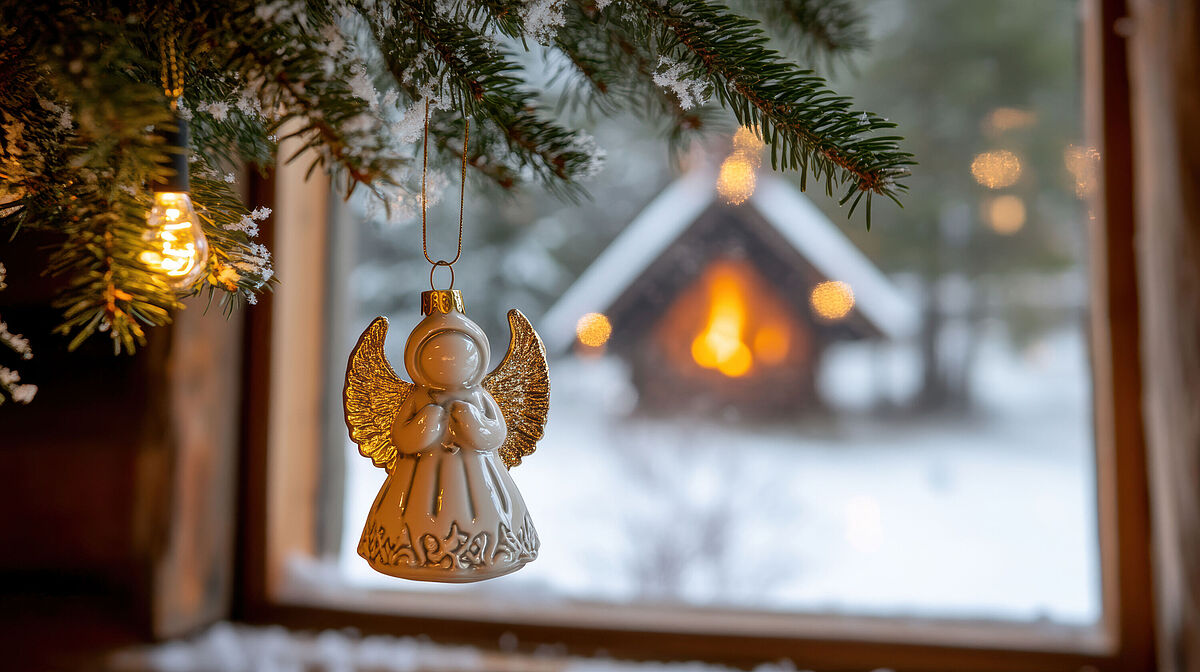 Engel im warmen Licht Ein Weihnachtsengel aus Ton hängt im Fesnter mit Tannenzweigen. Vor dem Fenster ist ein verschneiter Garten mit einer Hütte, die im warem Licht erstrahlt.