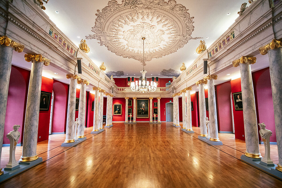 Die Aula der Universität Greifswald Prächtiger Barocksaal mit roten Wänden, Stuck an der Decke, an dem ein Kronleuchter hängt, weiß-marmorierte Säulen neben Büsten auf einem glatt gewienerten Holzfußboden.