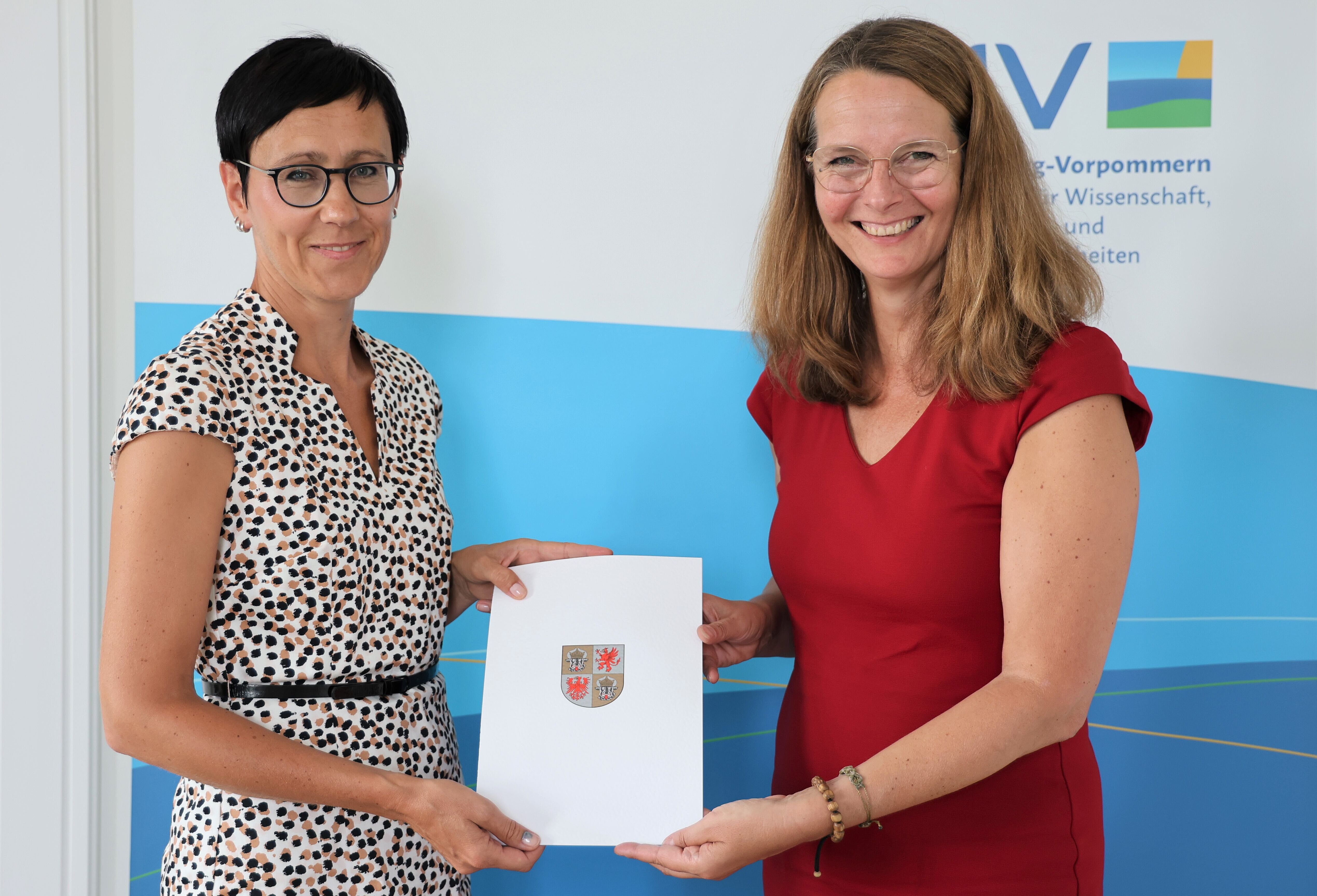Dr. Juliane Huwe und Bettina Martin Auf dem Bild sehen wir, wie Wissenschaftsministerin Bettina Martin die Ernennungsurkunde an Dr. Juliane Huwe übergibt. Beide halten die Urkunde in die Kamera.