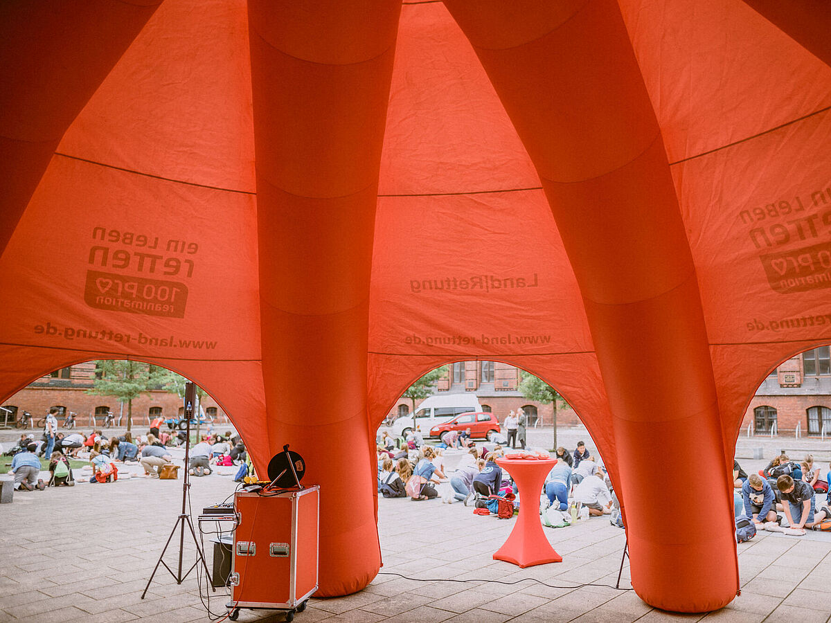Kinder- und JugendUNI Zu sehen ist ein großes orangefarbenes Zelt. Im Hintergrund knien viele Kinder während einer Rettungsübung zur Wiederbelebung von Menschen.