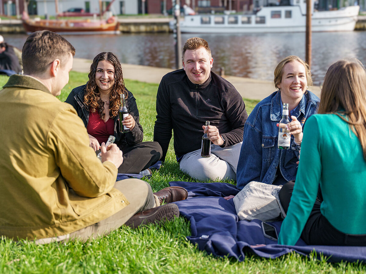 Studierende am Hafen Fünf Studierende sitzen auf dem Rasen am Ryck, unterhalten sich und halten Erfrischungsgetränke in der Hand.