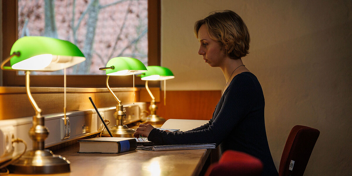 Symbolbild Philosophie Im Lesesaal einer Bibliothek liest eine Studentin in einem Buch. Auf den Tischen leuchten grüne Tischlampen.