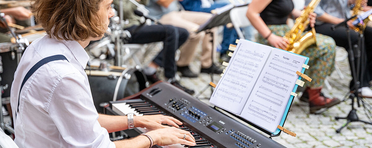 Dies Academicus 2024 Ein Mann sitzt spielend am E-Piano. Im Hintergrund sitzen Menschen mit Blasinstrumenten.
