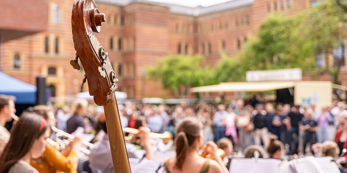 Konzert beim Dies Academicus Der Hals einer Violine ist im Vodergrund zu sehen. Im Hintergrund sitzen schemenhaft Menschen, die dem sommerlichen Konzert beiwohnen.