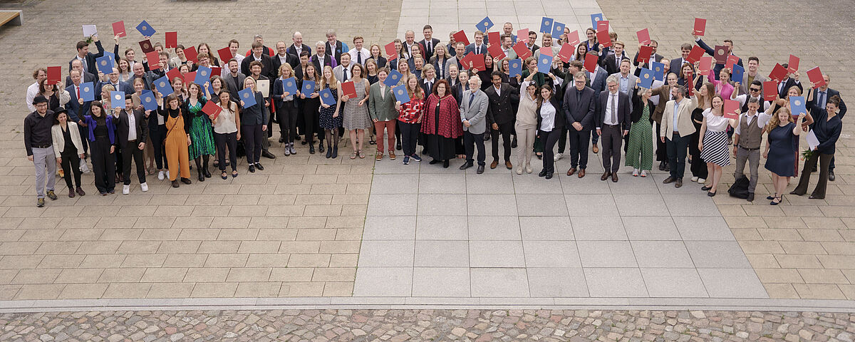 Verleihung akademishcer Grade Viele Menschen stehen für ein Gruppenbild im Innenhof einer alten Universität und halten Abschlussurkunden in die Luft.