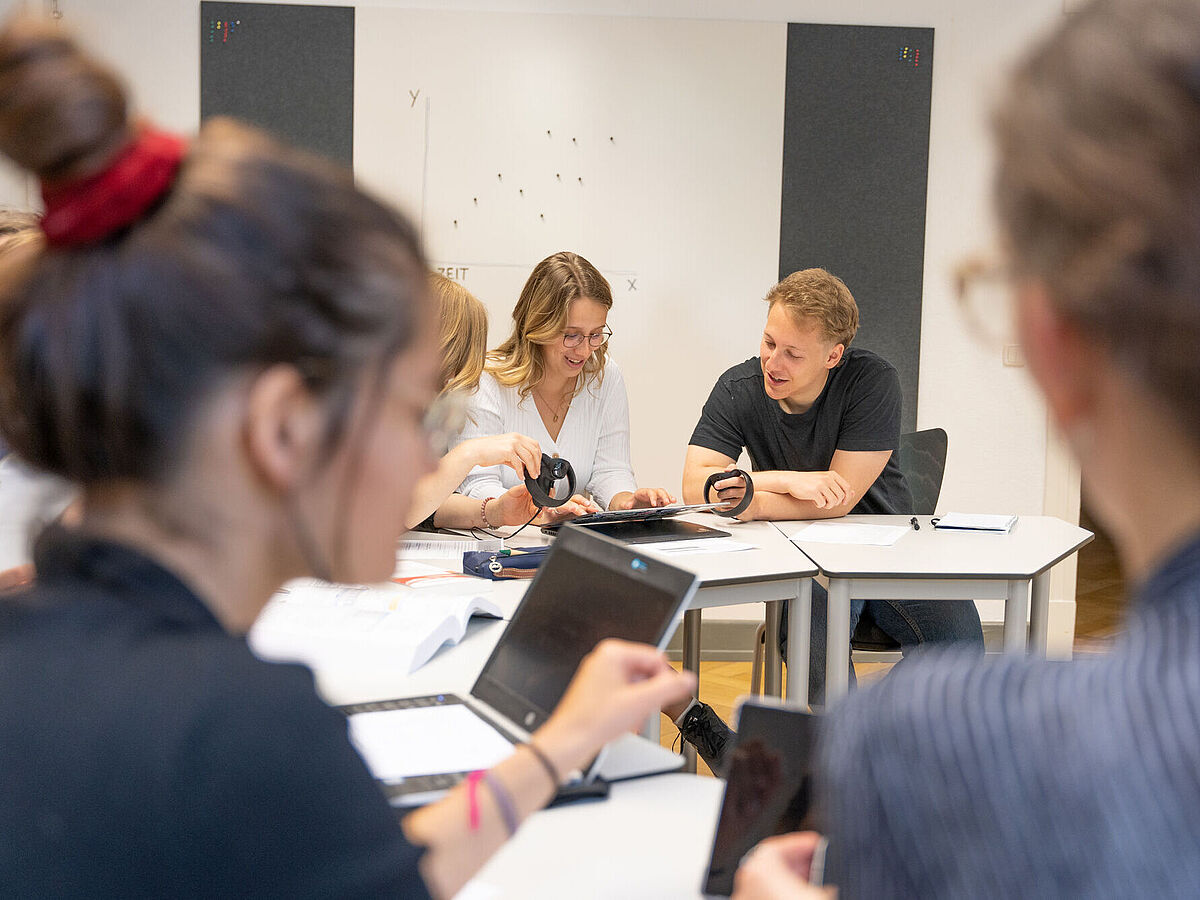 Symbolbild Meeting Im Meeting: Eine Gruppe von Menschen sitzt an weißen Tischen in einem Raum und arbeitet gemeinsam mit Tablets und VR-Controllern. Der Fokus liegt auf zwei Personen in der Mitte, die lächeln und ein Gerät bedienen.