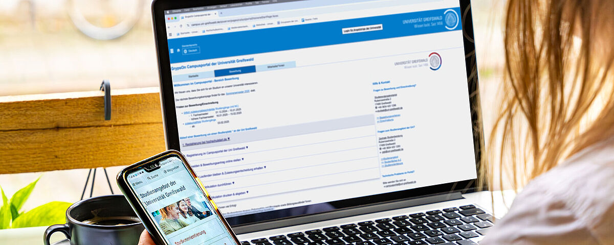 Symbolfoto Einschreibung Eine Person sitzt vor ihrem geöffneten Laptop und schaut zusätzlich in ihr Smartphone, wo die Seite zur Studienorientierung geöffnet ist.