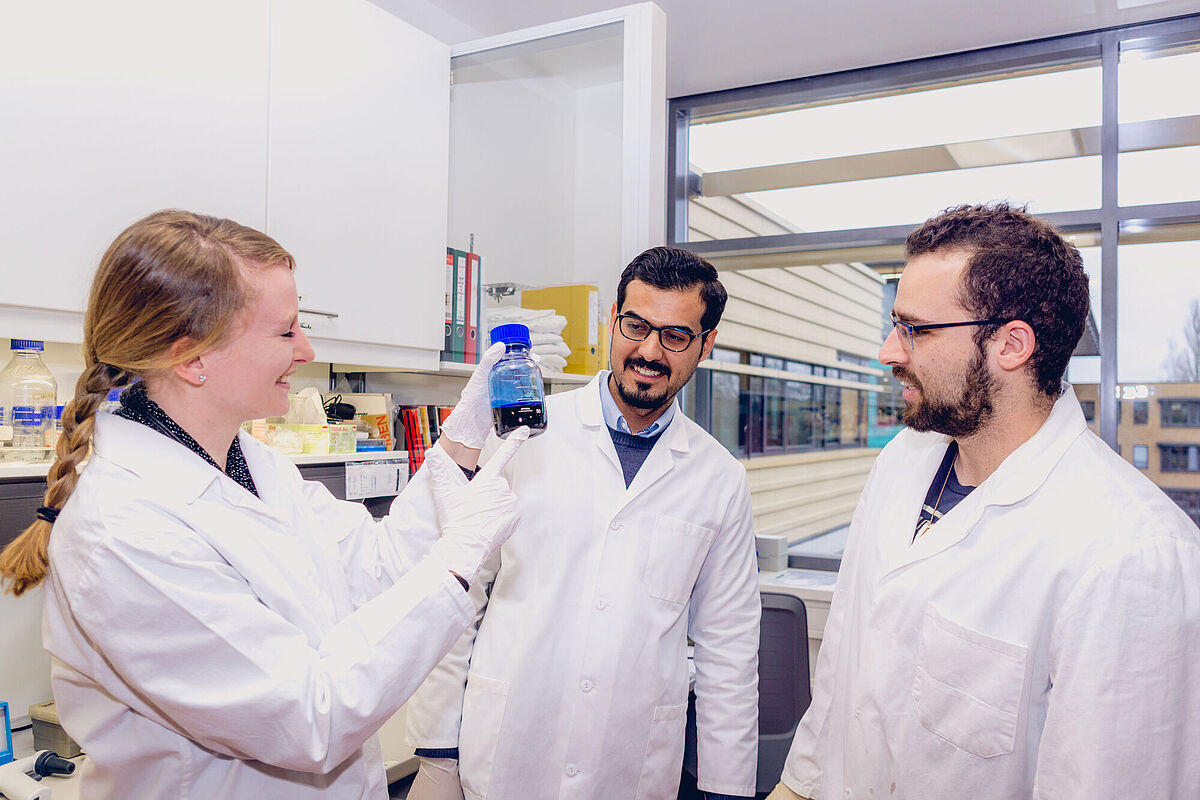 Symbolbild Forschung Drei Personen in weißen Laborkitteln betrachten in einem Labor eine blaue Flüssigkeit in einem Glasbehälter, die von einer Frau hochgehalten wird.
