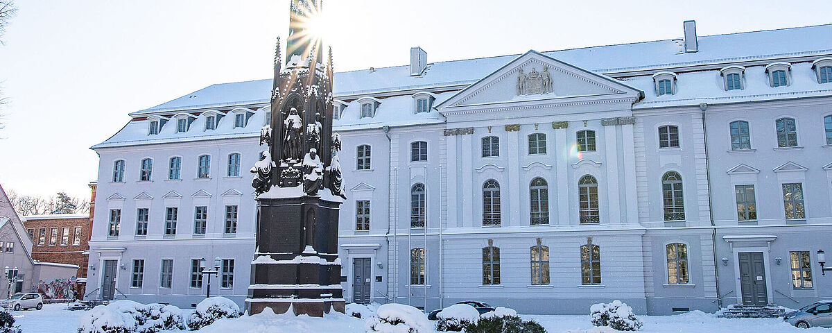 Winterbilder2021-LukasVoigt Das Hauptgebäude der Universität im Winter mit Schnee. Die Sonne scheint durchs Rubenowdenkmal.