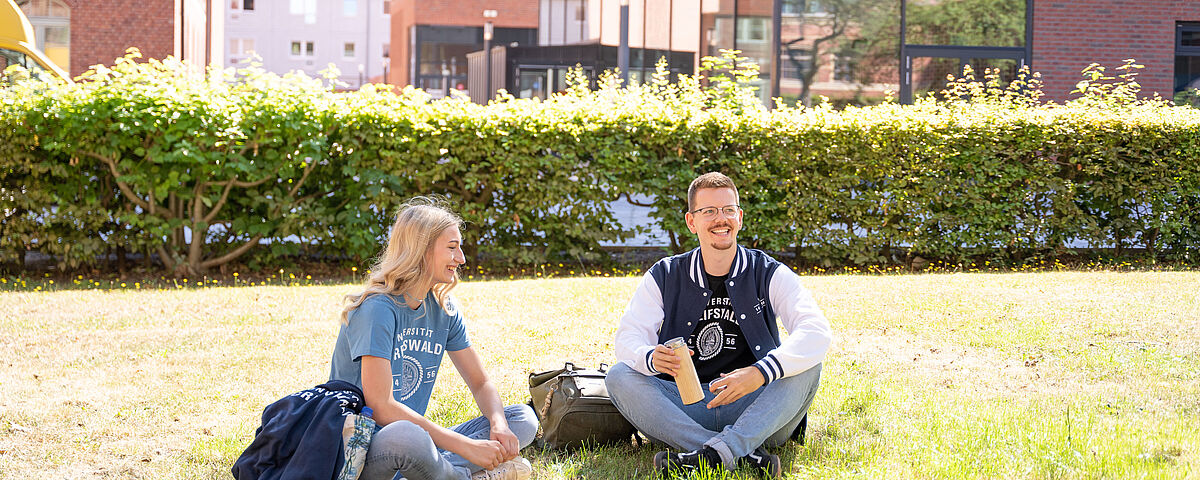 Uni Greifswald Merchandiseprodukte Eine Studentin und ein Student sitzen auf der sonnigen Wiese auf dem Lohmeyer-Campus, unterhalten sich und tragen dabei ein T-Shirt und eine Jacke mit dem Uni-Logo