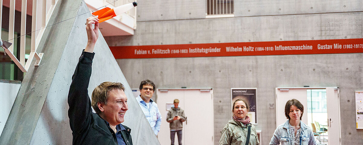 HIT 2025 Wissenschaftlicher Arbeiter aus dem Physik-Institut hält eine rot-durchsichtige "Rakete", die an einer nach oben ragenden Leine befestigt ist. Er lächelt. Um ihn herum stehen mehrere Zuschauende.