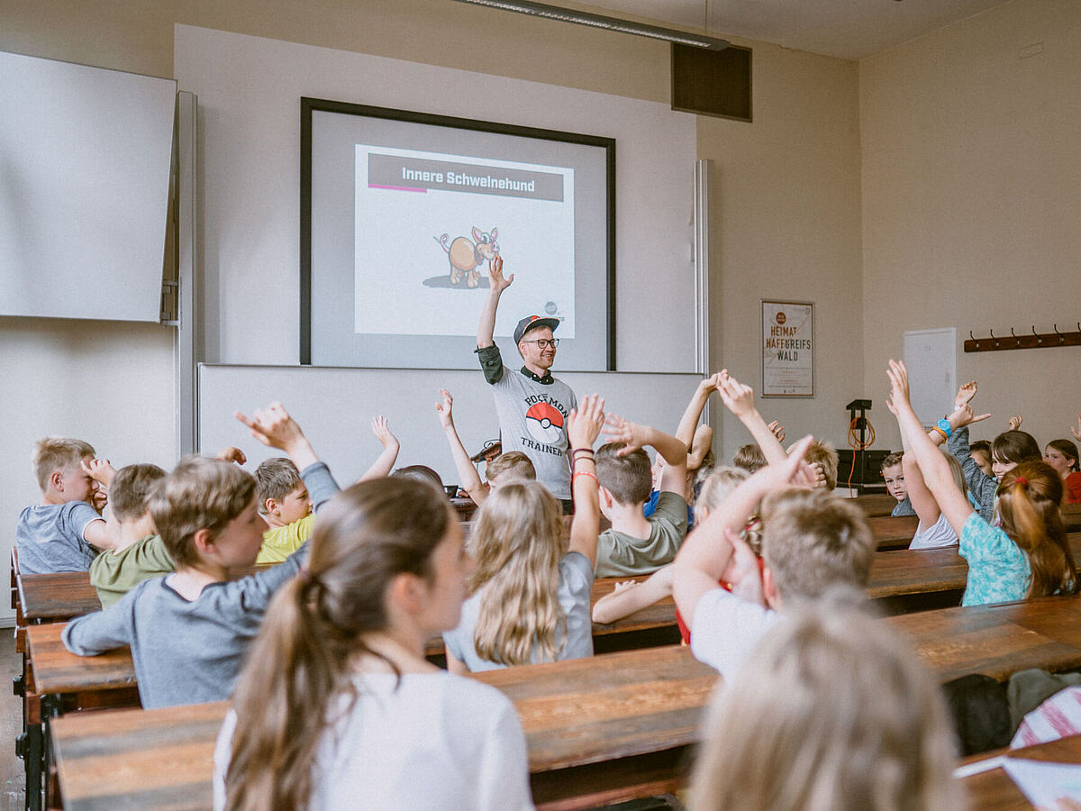 Ein Hörsaal mit Kindern, die begeistert ihre Hände heben, während ein Mann vorne steht und auf eine humorvolle Präsentation mit der Überschrift „Innerer Schweinehund“ zeigt.