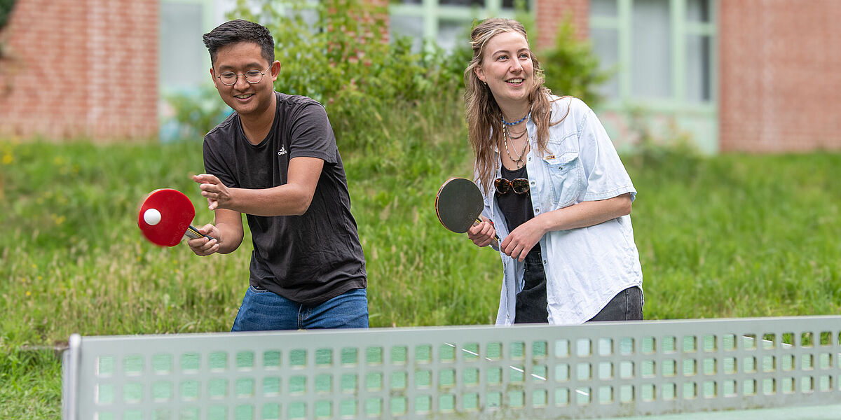 Tischtennis am Wall Ein Student und eine Studentin stehen hinter einer Tischtennisplatte, bereit für die Ballannahme.