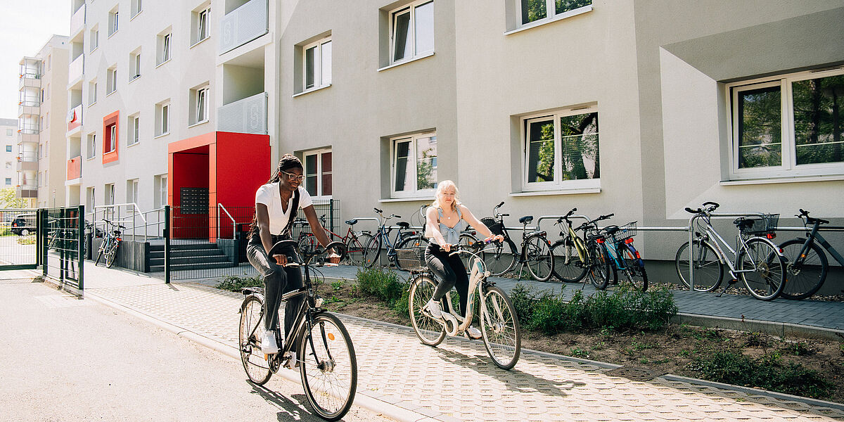Wohnheim Makarenkostraße Zwei Studentinnen fahren mit ihrem Fahrrad bei Sonnenschein vor einem grau-weiß gestrichenem Wohnheim vorbei