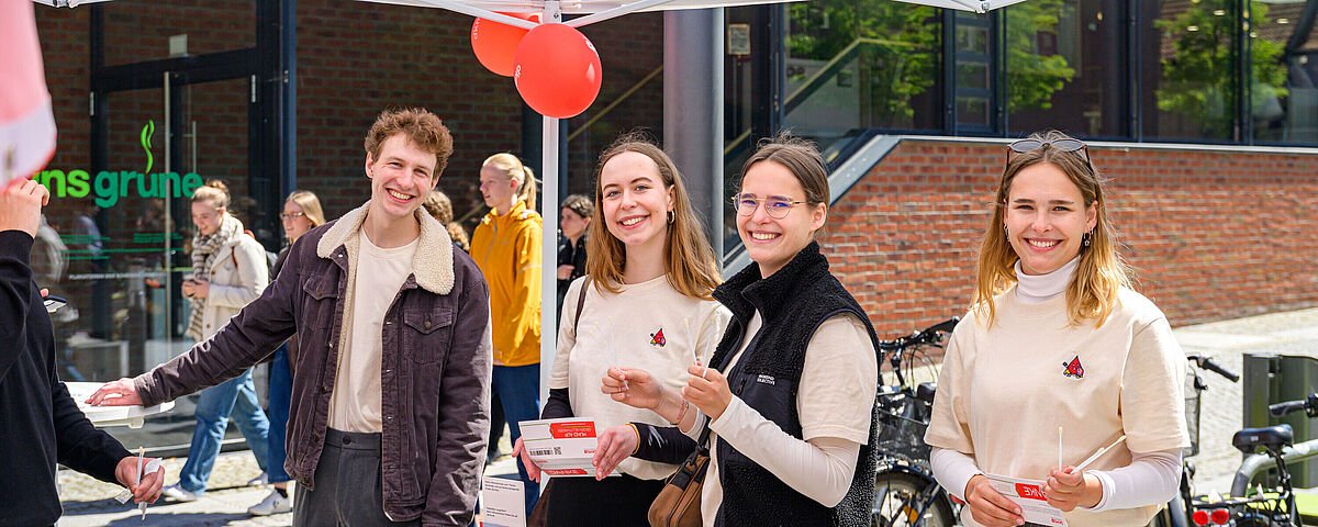 HIT 2025 DKMS Vier Studierende stehen unter einem weißen Zelt vor der Cafeteria "ins grüne" am Campus Loefflerstraße. Sie halten Infomaterial von der DKMs in der Hand und lächeln in die Kamera.