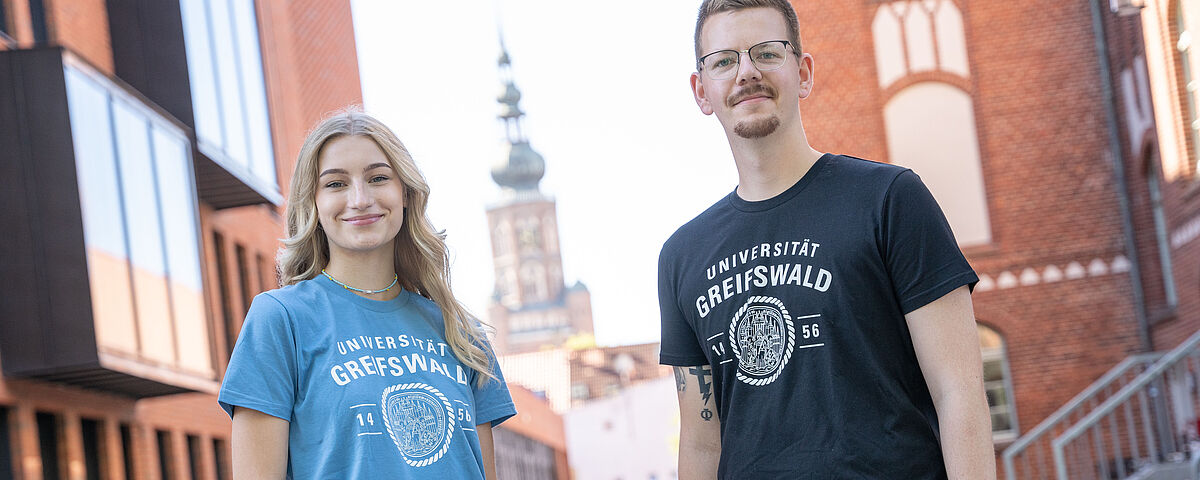 Uniladen T-Shirts Eine Studentin und ein Student stehen vor dem Campus in Backsteinoptik und tragen jeweils ein T-Shirt mit Uni-Logo