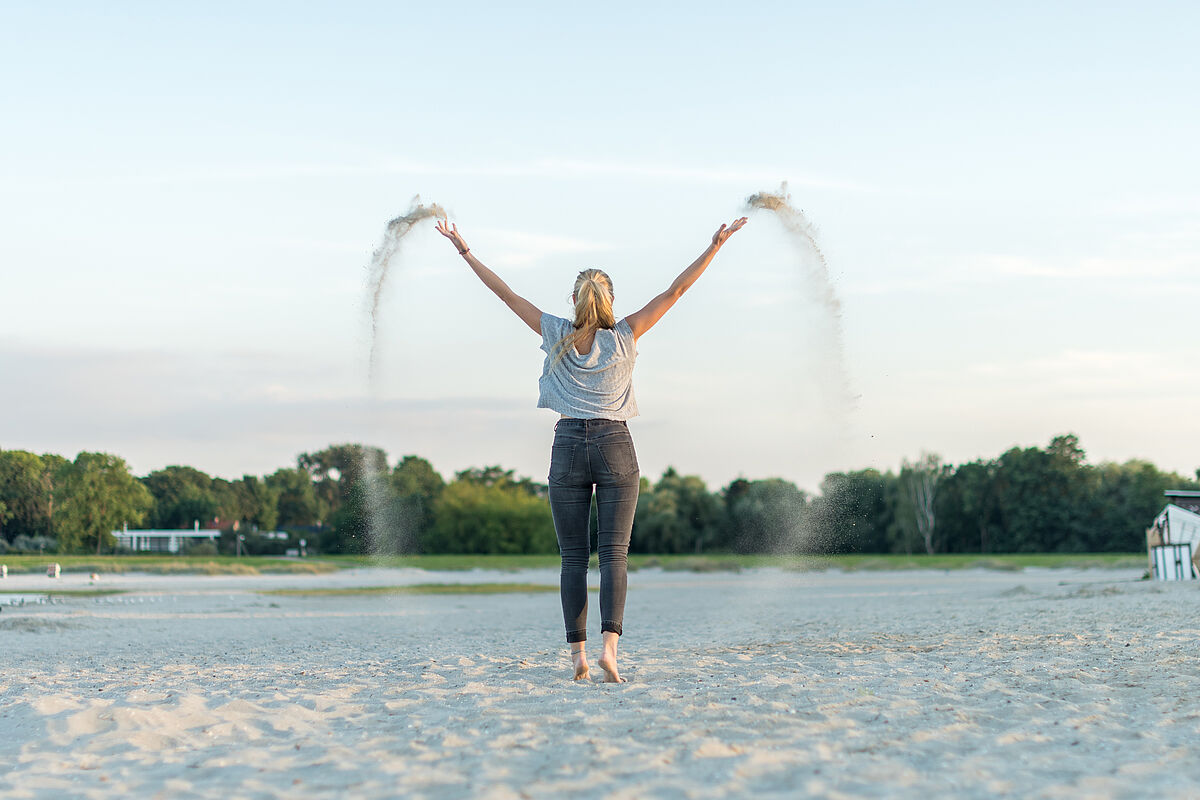 Im Strandbad Eldena Eine Frau steht mit dem Rücken zum Betrachter barfuß auf dem Sand des Strandbad Eldena. Sie schwingt die Arme nach oben und verstreut Sand
