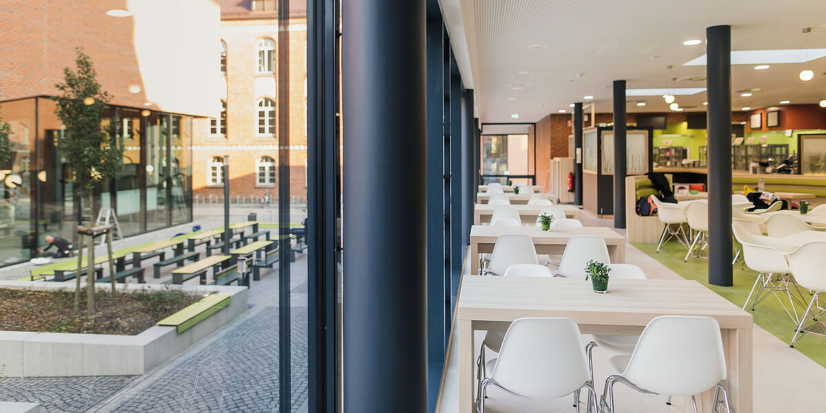 Café "ins grüne" am Campus Loefflerstraße Ein heller, freundlicher Innenraum mit weißen Stühlen und hellen Tischen. Links der Blick auf den Innenhof des Backstein-Campus.