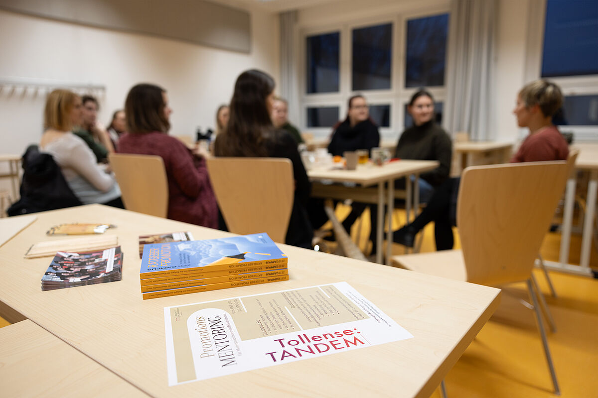 Netzwerktreffen. Im Hintergrund Frauen am Tisch im Austausch. Im Vordergrund sieht man einne Tisch mit Flyern, unter anderem vom Programm Tollense:TANDEM.