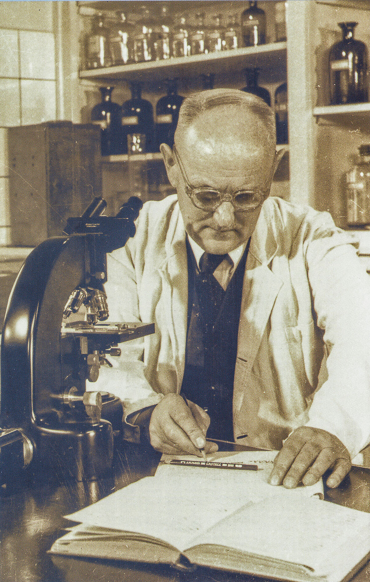 Prof. Dr. Gerhard Domagk beim Eintragen mikroskopischer Befunde. Foto: Gerhard Domagk. Ein Mann im Laborkittel beim Eintragen mikroskopischer Befunde in eine Laborjournal. Neben ihm steht links ein Mikroskop.