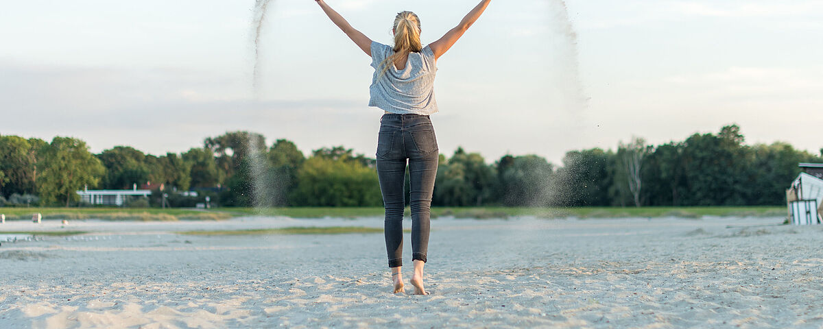 Im Strandbad Eldena Eine Frau steht mit dem Rücken zum Betrachter barfuß auf dem Sand des Strandbad Eldena. Sie schwingt die Arme nach oben und verstreut Sand