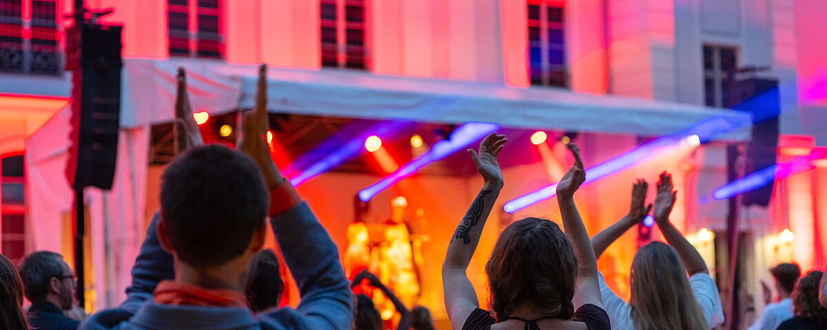 Symbolbild Party Mehrere Menschen von hinten heben ihr Hände vor einer blau, orange und pink beleuchteten Konzertbühne.