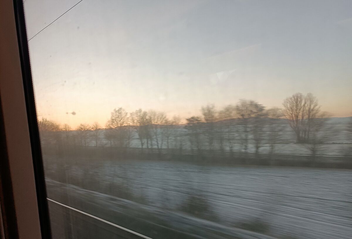 Foto aus dem Zugfenster heraus. Draußen liegt Schnee und es ist kurz vor dem Sonnenuntergang.