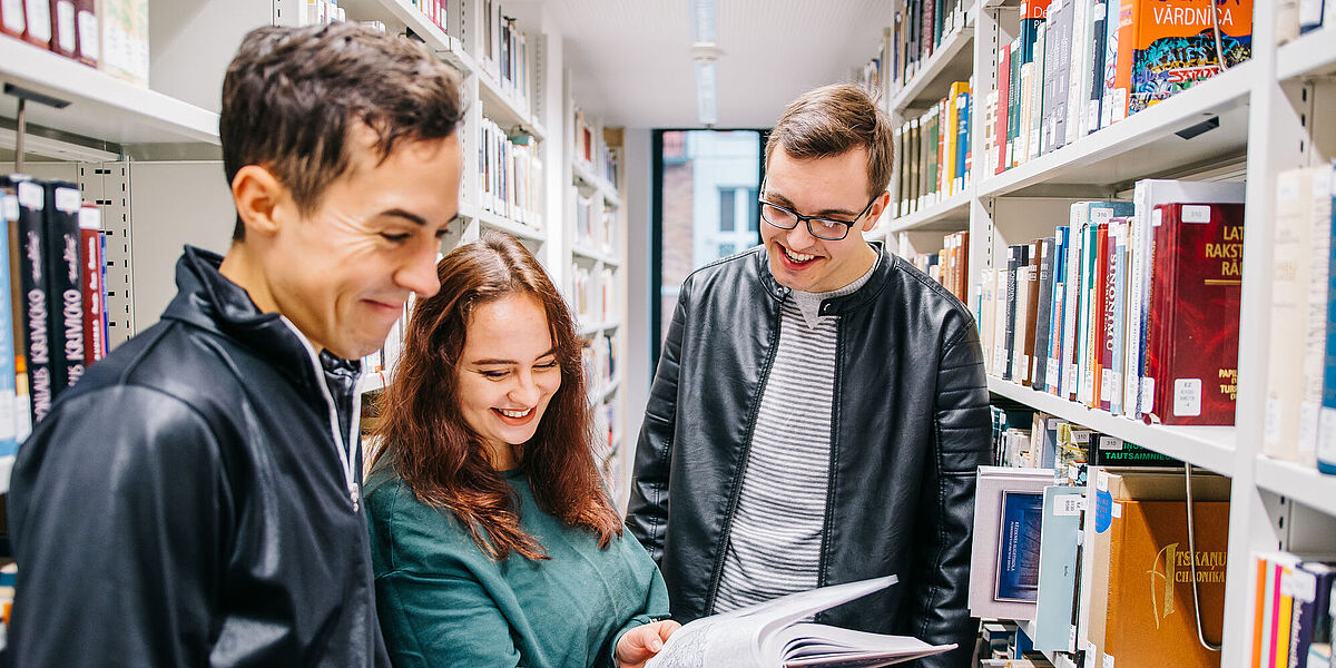 Symbolbild Bibliothek Drei Personen stehen in der Bibliothek zwischen hohen Regalreihen und lesen in einem Buch.
