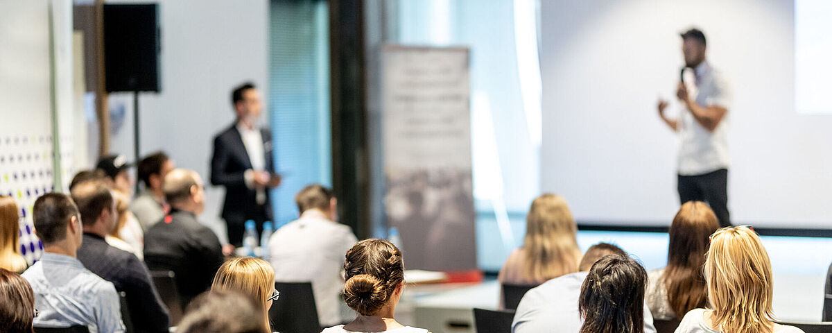 Symbolbild Vortrag Auf dem Bild ist ein Redner auf einer kleinen Bühne mit einem Moderator und Zuschauern zu sehen.