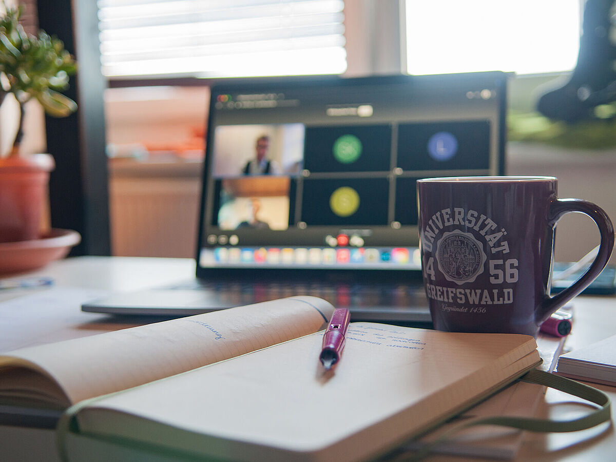 Symbolbild Videokonferenz Ein Schreibtisch mit einem aufgeklappten Laptop, auf dem ein Videocall läuft, einer Tasse mit Universitätslogo und einem offenen Notizbuch mit Stift.