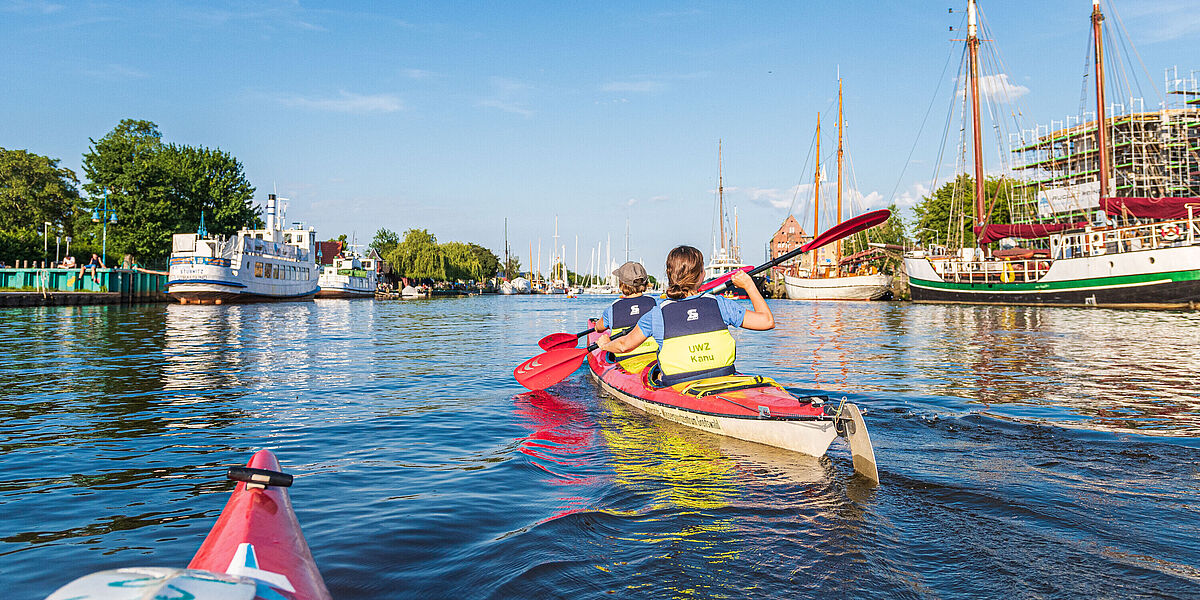 Kanukurs auf dem Ryck Zwei Studierende sitzen in einem Kanu und paddeln bei sonnigem Wetter auf dem Ryck mit Blick auf den Museumshafen.