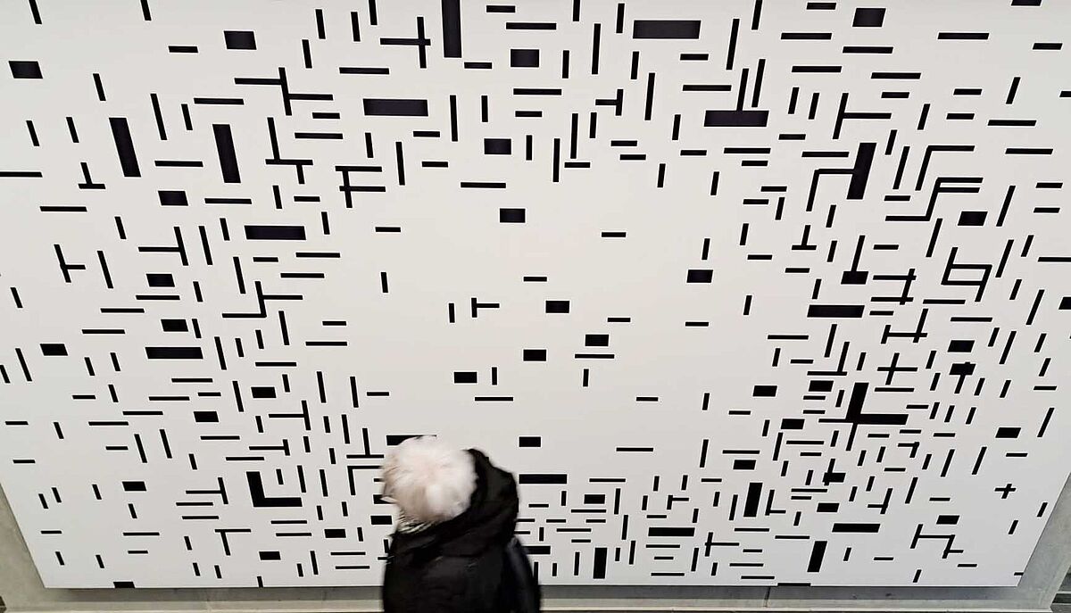 Man sieht ein Kunstwerk von Susanne Gabler, ein großes Bild mit vielen unterschiedlich großen und verschieden geformten schwarzen Rechtecken auf weißem Hitergrund. Sie sind ungleichmäßig auf dem Bild verteilt, sodass es so wirkt, als ergäben sie einen sich zu den Rändern verlaufenden Kreis.Davor der Kopf einer besuchenden Person, die das Werk betrachtet.